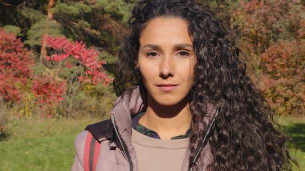 Közelkép női portré komoly fiatal gyönyörű spanyol túrázó lakókocsi modell göndör haj nézi a kamera kint áll a természetben pózol ősszel erdő mosolygós utazás hátizsák — Stock videók
