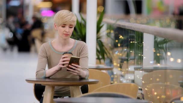 Middelbare leeftijd volwassen jaren 40 Kaukasische vrouw zitten aan cafe tafel het drinken van koffie thee uit papieren beker op zoek in mobiele telefoon typen smartphone chatten browsen kopen online app met behulp van wi-fi sms-en — Stockvideo