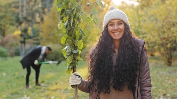 Detailní záběr mladá šťastná usměvavá dívka stojící venku držení sazenice v ruce muž kopání otvor pro výsadbu stromu v pozadí dobrovolníci eko aktivisté chránit přírodu zeleň planety zlepšení ekologie — Stock video