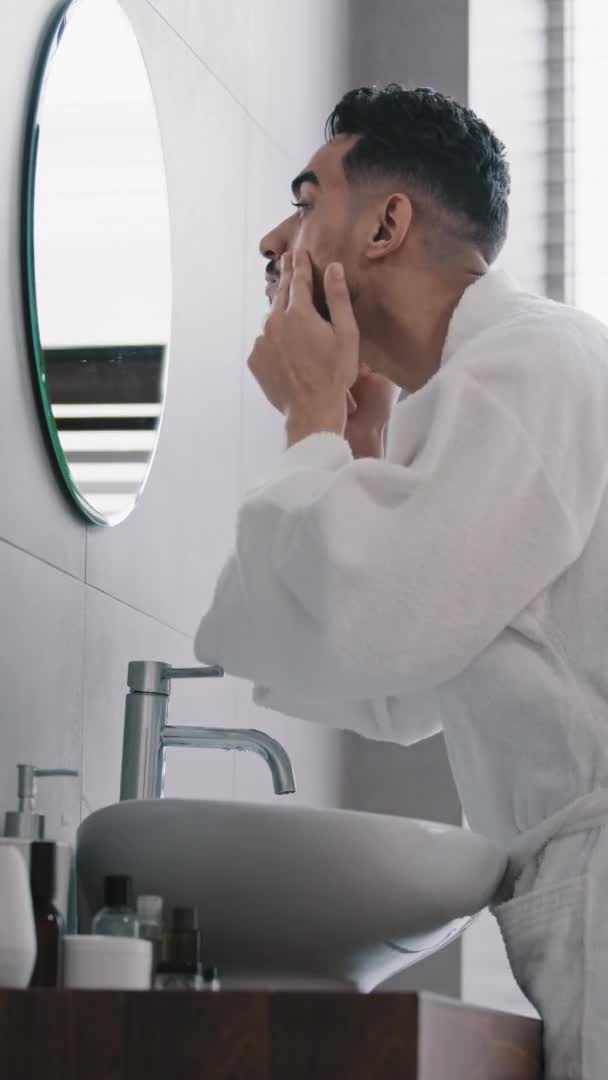 Vue verticale arabe indien arabe barbu hispanique homme en peignoir appliquer crème hydratante sur la peau du visage regarder dans le miroir dans la salle de bain montrant pouce vers le haut heureux succès soins de la peau matin beauté faciale — Video