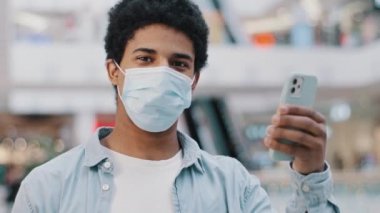 Koruyucu tıbbi maskeli Afrikalı adam kameraya destek jesti gösteren akıllı telefonuna bakıyor. Baş parmaklar gibi. Tıbbi uygulama uzaktan doktorluk hizmeti öneriyor.