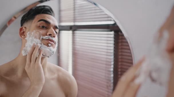 Hispano hombre barbudo árabe árabe indio milenial 30s modelo de hombre utilizando espuma blanca gel de afeitar cosméticos naturales frota las mejillas de la barba baño mirando el espejo. Guy prepararse para afeitarse bigote barba — Vídeos de Stock