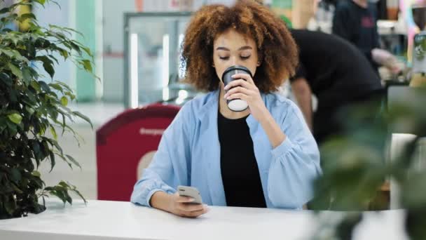 Africana 20s mujer multirracial biracial dama con corte de pelo rizado sentado en la mesa de café beber té de café de la taza de papel mirando en el teléfono móvil mecanografía teléfono inteligente chat navegación comprar aplicación en línea — Vídeo de stock