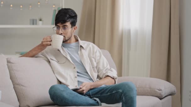 Тысячелетний арабский парень индиец красивый 30-летний мужчина сидит дома на диване и пьет горячий чай капучино какао из чашки получает сообщение глядя на телефон улыбаясь болтая с друзьями со смартфоном — стоковое видео