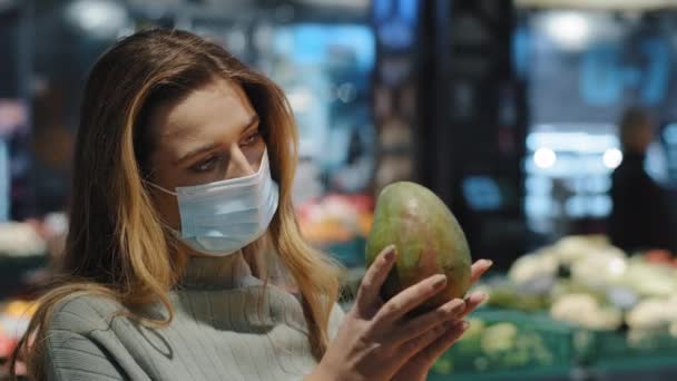 Καυκάσιος κορίτσι αγοραστής 30 γυναίκα στο ιατρικό πρόσωπο προστατευτική μάσκα κρατώντας πράσινο μάνγκο τροπικά φρούτα στο κατάστημα ψώνια στο μπακάλικο βιταμίνη διατροφή υγιεινό τρόπο ζωής χορτοφαγία οικολογική έννοια — Αρχείο Βίντεο