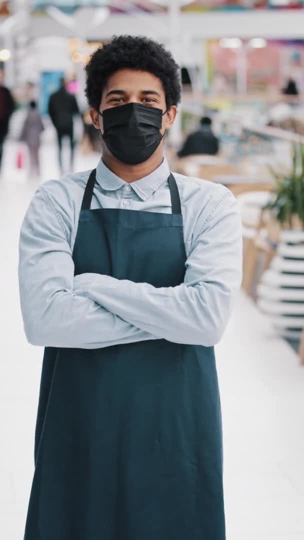Vertikální pohled africký americký kavárna pracovník multiracial člověk chlap restaurace muž prodejce v ochranné lékařské masky při pohledu kamery představuje křížení zbraní číšník prodejce v zástěře pózování s zkříženýma rukama — Stock video