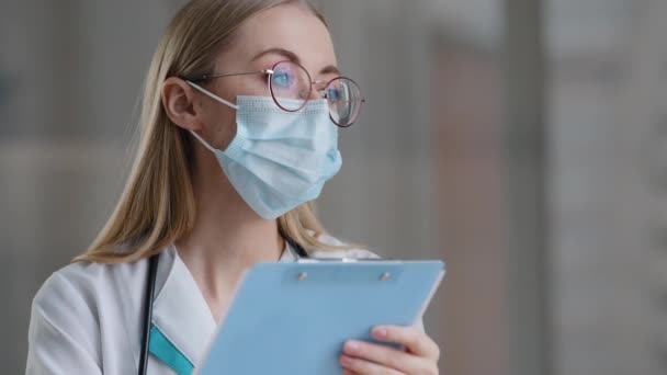 Retrato caucásico mujer médico enfermera en médico protector máscara escritura información en médico registro diario femenino en gafas terapeuta trabajo clínica hospital escritura notas cita — Vídeo de stock