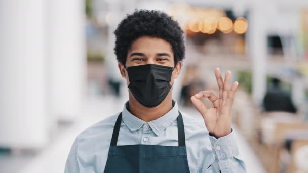 Gemaskerd Afrikaans Amerikaans man cafe werknemer ober verkoper in schort medisch gezichtsmasker succesvolle man kleine ondernemer op zoek camera shows ok eens prima goed teken groot gebaar pandemie regels — Stockvideo