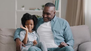 Afrikalı Amerikalı bir aile kanepeye oturmuş el sallıyor yetişkin bir babaya merhaba diyor kameraya sarılıyor, gülümsüyor dişler sağlıklı beyaz dişler gösteriyor video görüntülü kamera görüntüsü.