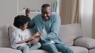 Mutlu Afro-Amerikan aile yetişkin baba kızıyla oynuyor oturma odasında kanepede oturuyor babası gıdıklıyor sevimli kızı gıdıklıyor ve gülüyor birlikte vakit geçirip eğleniyor.