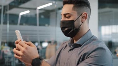 Gömlekli çok ırklı Arap adam ofiste tıbbi yüz koruma maskesi takıyor cep telefonu işadamı ile mesajlaşıyor akıllı telefonlu ücretsiz internet üzerinden iş mesajı kullanıyor şirkette.
