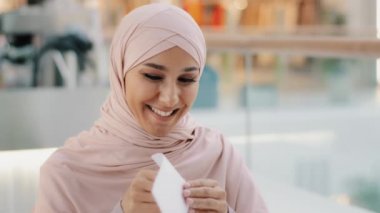 Yakın plan, heyecanlı, tesettürlü, tesettürlü, mutlu bir Arap kadın, harika haberler okuyor, gülümsüyor, tıbbi test sonuçları, yeni iş maaşı için onaylanıyor.