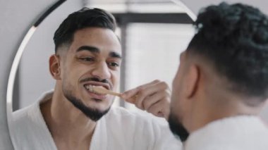 Banyoda aynada yansıyan erkek yansıması. Sakallı, sakallı, sağlıklı dişlerini beyazlatıcı macunla fırçalayan erkek. Evde sabah temizliğinde diş fırçası kullanıyor.