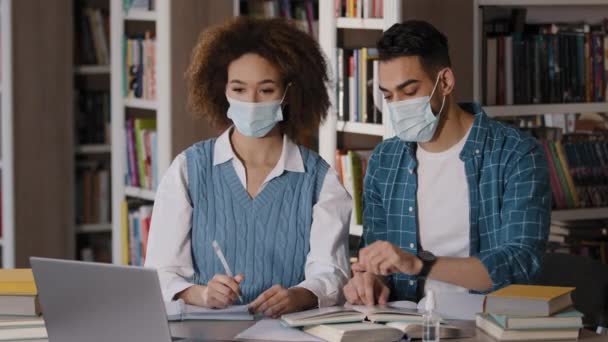 Dois estudantes menina e cara em máscaras protetoras sentado na mesa ouvindo palestra on-line de laptop aprendizagem à distância à procura de informações no livro didático virar notas de escrita página no caderno — Vídeo de Stock