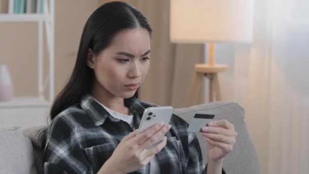 Angry azjatyckie kobieta siedzieć na kanapie w domu hold karta kredytowa za pomocą telefonu komórkowego spróbuj zapłacić z e-bank app problemy z płatnościami błąd bankowy błąd nie pieniądze online operacja finansowa nieudana transakcja — Wideo stockowe