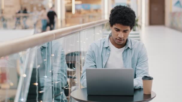African American επικεντρώθηκε σοβαρή χιλιετή επιχειρηματίας άνδρας ελεύθερος επαγγελματίας φοιτητής φοιτητής τύπος χρήστη κάθεται στο τραπέζι στο καφέ με καφέ εργασίας laptop πληκτρολογώντας περιήγηση e-learning μελέτη net wifi web ecommerce — Αρχείο Βίντεο