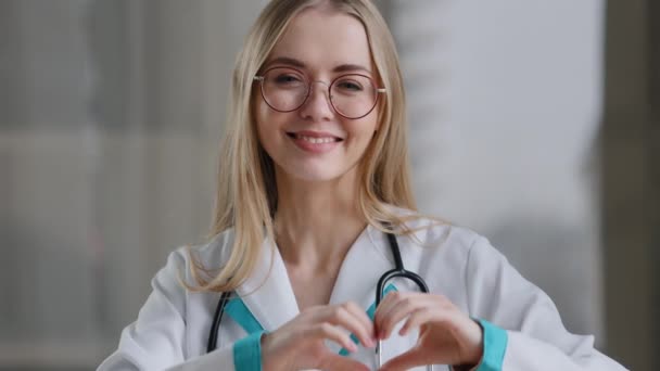 Kaukázusi orvos szakorvos nővér nő gyakorló terapeuta szemüvegben a klinikán az asztalnál mutatja szív alakú jele irgalom szeretet kedvesség segítségnyújtás gyógyszer segít nemzetközi orvosi nap — Stock videók