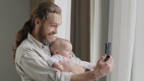Šťastný úsměv kavkazský otec dospělý tatínek drží novorozeně dítě doma stojí v blízkosti okna tatínek s malou dcerou syn novorozenec tvorby fotografie video vlog on-line proud konference vzdálený hovor — Stock video