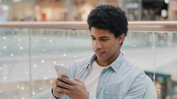 Afrikalı odaklı milenyum adamı, kapalı mekanda oturup mobil sohbet uygulamasını araklıyor online alışveriş oyunu, akıllı telefon sohbetleri, ücretsiz Wi-Fi kullanıcı sohbet sosyal medya. — Stok video