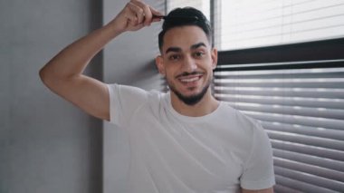 Arap İspanyol bir adam kameraya bakıyor saç fırçasıyla saçını tarıyor iş günü için hazırlanıyor beyaz tişörtlü, sabah güzelliğinde, saç kesiminde, randevuya hazırlanıyor.