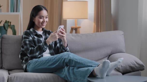 ソーシャルメディアでリラックスしたニュースフィードを見て自宅でソファに座っているアジアの女の子オンラインショップインターネットストアを選択します日付サイトのWi-Fiスマートフォンのショッピングポストストーリースワイプタッチスクリーン — ストック動画