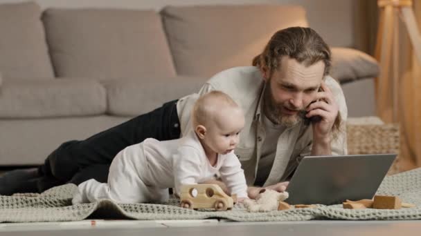 Biały, wielozadaniowy ojciec pracujący w domu z kwarantanną dla laptopa z niemowlęciem leżącym na podłodze i mówiącym przez telefon rozwiązującym problemy biznesowe. Wyczerpany rodzic z nadpobudliwym dzieckiem — Wideo stockowe