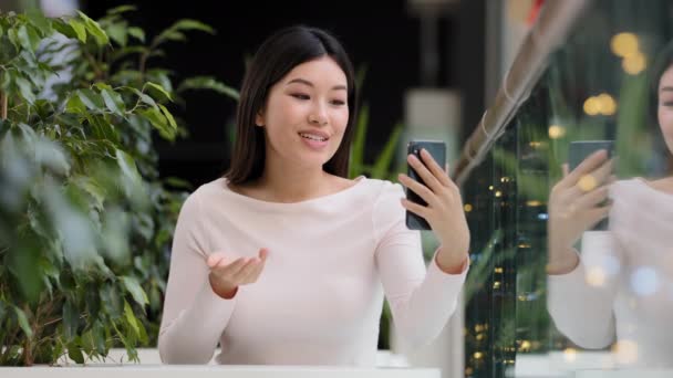 Aziatisch koreaans vriendschappelijk meisje japans dame chinees vrouw glimlachen zwaaien hallo naar telefoon webcam vrouw met mobiele gadget praten maken video bellen in cafe office tafel chatten door virtuele conferentie app — Stockvideo