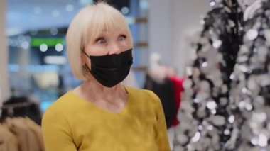 Medikal maskeli orta yaşlı bir kadın giyim mağazasındaki müşteriler abartılı, parlak, parıltılı elbiseye hayranlıkla gülümseyen, parıltılı kıyafetlerle oynayan müşterilere şaşırmış bir şekilde bakıyorlar.