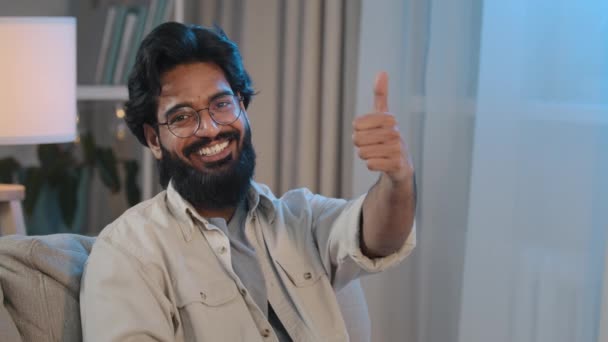 Retrato feliz satisfeito alegre árabe indiano homem barbudo masculino em óculos sentado em casa em novo apartamento no sofá confortável olhando para a câmera sorrindo mostrando polegares para cima recomenda agência imobiliária — Vídeo de Stock
