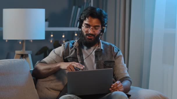 Árabe indiano engraçado homem barbudo feliz usa fones de ouvido sem fio se divertindo ouvindo música com laptop em casa sentado sofá noite tarde tempo escuridão quarto relaxante canto música desfrutar de áudio app — Vídeo de Stock