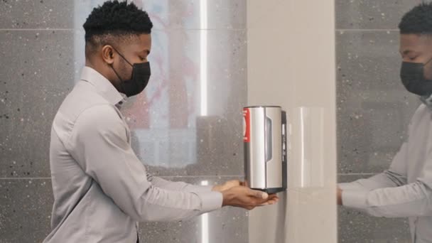 Young african american man in protective mask stands in public toilet dries hands under jet of hot air using hand dryer surprised by modern technology touchless washing hygiene and sanitation concept — ストック動画