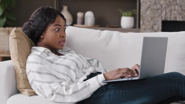 African girl freelancer american biracial woman lying on couch at home looking in laptop reading unexpected bad news gossip receiving notification opens mouth wide in surprise shock looking to camera — Stock Video