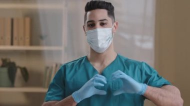 Portreci genç Arap doktor hemşire yüz maskesi üniforması giyiyor. Kamera karşısında aşk işareti. Eldiven giymiş erkek eli.