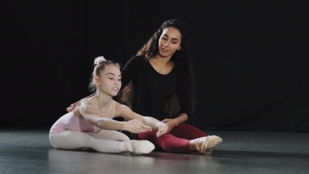 Adult woman coach dancer teacher sitting on floor with girl teenager child student acrobat beginner ballerina gymnast leaning down flexing body coach helps with stretching advice trains ballet class — Stock Video