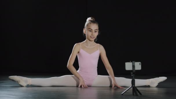 Mädchen Teenager Kind Ballerina Akrobat Turnerin sitzt auf Boden Tanzkurs Online-Lektion mit Internet-Trainer Lehrer Fernbedienung mit Smartphone auf Stativ sitzt auf Bindfäden Aufzeichnung Video-Blog vlog — Stockvideo
