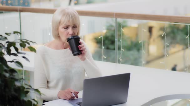 Mature caucasian serious woman office worker businesswoman sitting at desk looking in screen laptop drinking hot coffee from disposable cup writes notes makes plan studies new information checks mail — ストック動画