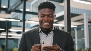 Denetmen Afrikalı erkek ofis yöneticisi elinde akıllı telefonu tutarak ekranda kaydırma sayfasına dokunuyor. İnternet içeriğinde geziniyor, gülüyor ve aletin üzerinde işaret ediyor.