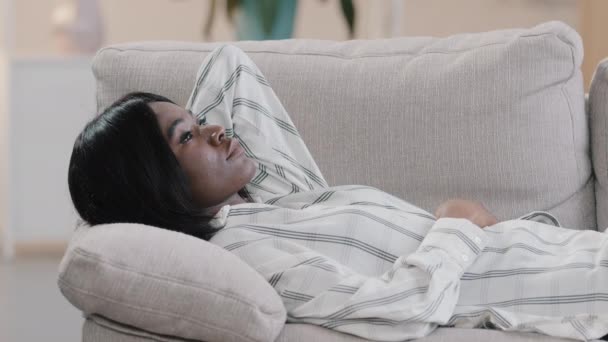 Jeune femme américaine africaine rêveuse calme et dispendieuse reposant sur un canapé confortable dans le salon, jouissant d'une sieste relaxante, d'une sieste saine et paresseuse, d'une sieste diurne, d'une rêverie, d'un calme, d'une main derrière la tête. — Video