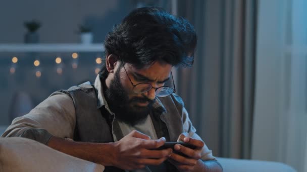 Adulte arabe homme barbu indien avec des lunettes est assis sur le canapé à la maison le soir dans la nuit dans la nuit dans l'obscurité tard jouer à des jeux mobiles en ligne compétition sur les gains de téléphone faire un geste de victoire addiction concept — Video