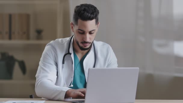 Homem concentrado sério médico usar casaco branco usando computador internet digitando nomeação no armário do hospital médico profissional masculino consultar paciente distantemente on-line bate-papo sentar na mesa — Vídeo de Stock