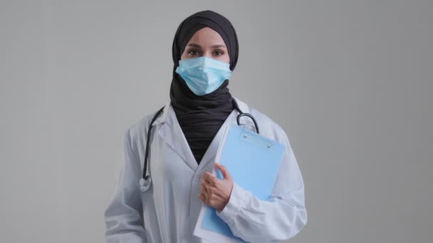 Портрет арабки-мусульманки женщины-врача-хирурга медсестры в хиджабе медицинской маске, позирующей со стетоскопом, держащей папку с документом, глядя в камеру, предотвращает коронавирусную концепцию — стоковое видео