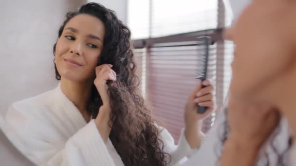 Close-up happy smiling young woman in bathrobe looking in mirror brushing combing long curly hair doing hairstyle daily care in bathroom in morning getting ready enjoying everyday routine at home — ストック動画