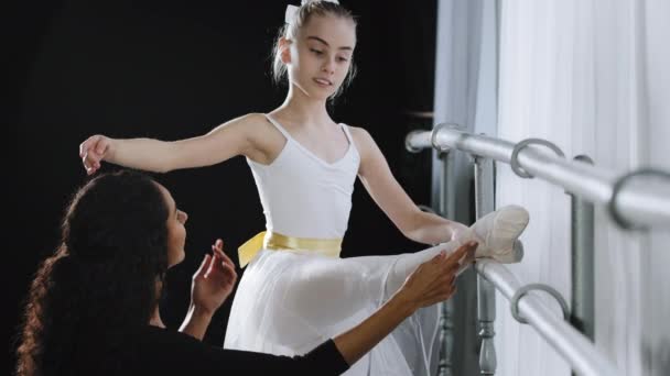 女青少年芭蕾舞女学生在芭蕾理发店附近做传统的运动听女教师的建议专业女教练帮助伸展腿保持童腿矫正姿势 — 图库视频影像