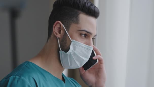 Λατίνος ιατρικός βοηθός γιατρού σε ιατρική μάσκα συμβουλεύεται ασθενή απομακρυσμένη συζήτηση θέματα από το τηλέφωνο ισπανόφωνος σύμβουλος χρησιμοποιώντας κινητό εφαρμογή υγειονομικής περίθαλψης στέκεται κοντά παράθυρο μακρινή έννοια διαβούλευση — Αρχείο Βίντεο