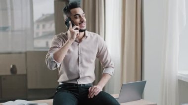 Gülümseyen Arap adam rahat erkek girişimci işadamı çalışma alanının tepesinde oturmuş müşteriyle cep telefonuyla görüşme yapıyor.