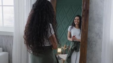 Komik, mutlu, genç, güzel bir kadın aynanın önünde neşeli bir şekilde dans ediyor. Dinamik hareket ediyor. Uzun kıvırcık saçlarla eğleniyor.