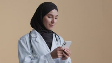 Doktor kadın terapist kadın tesettür beyaz üniforması giyiyor. Klinikte ayakta duruyor. Akıllı telefon mobil sağlık hizmetleri başvurusu yapıyor.