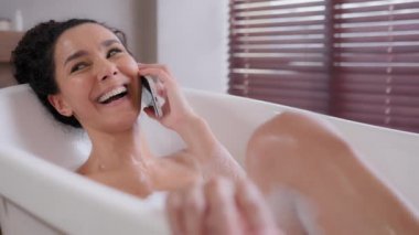 Genç, mutlu bir kadın köpük banyosunda uzanıyor banyoda dinleniyor cep telefonuyla konuşuyor gülüyor arkadaşça konuşmalar yapıyor akıllı telefon hijyenik günlük rutinini kullanarak hoş sohbetler ediyor.