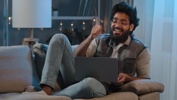 Excited arabian indian bearded man winner look at laptop celebrate success online victory get great opportunity sitting at sofa at home evening night dark read good news new job offer reach concept — Video