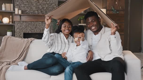 African family sit on sofa under carton roof. Homeowner father mother with child daughter at new house living room celebrate relocation day to own rented apartment kid girl holding a bunch of keys — ストック動画